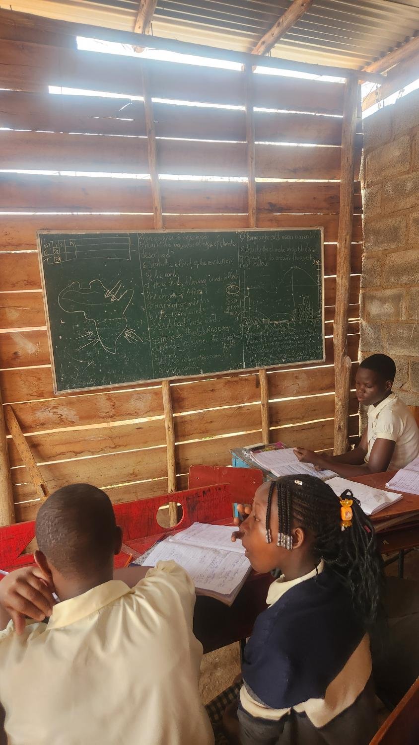 Pupils at desks under a timber and iron roof, with a geography lesson and a map of Africa on the board