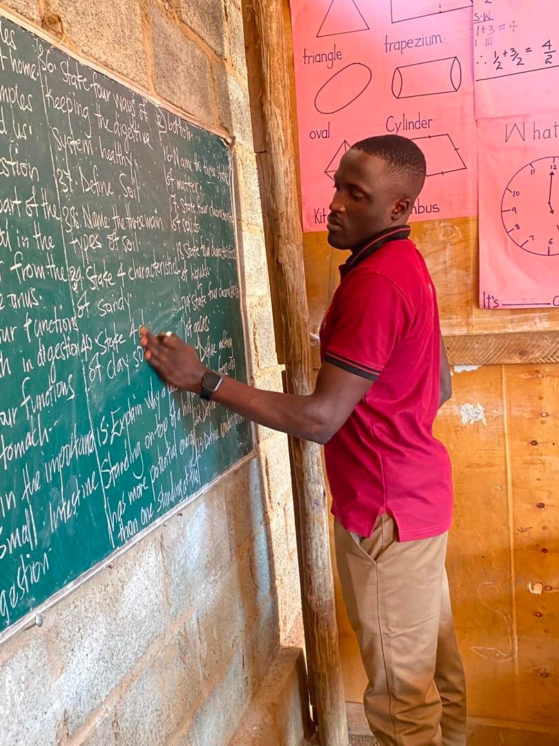 A teacher in a red shirt writes on a green chalkboard in a timber classroom