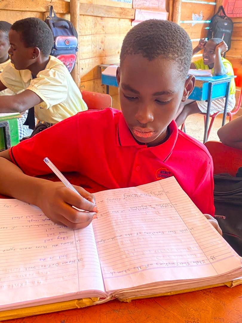 A boy in a red Mercy School shirt writes in an open exercise book