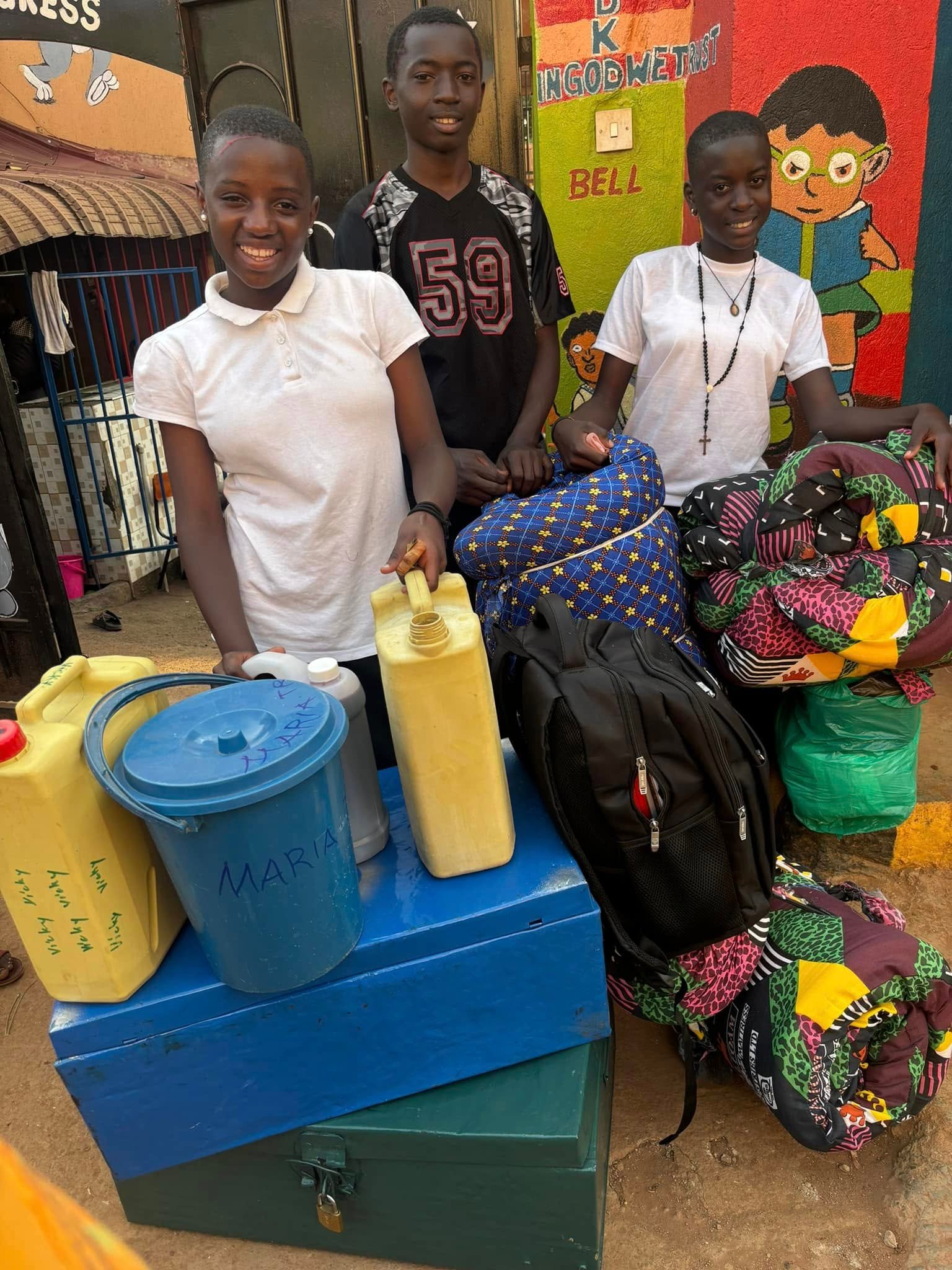 Three children stand with yellow water jerrycans, a blue bucket labelled Maria, backpacks and rolled bedding